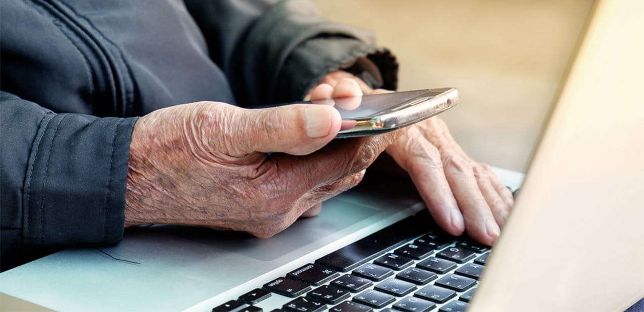 Close up of the hands of an elderly person working on a laptop and holding a cell phone.