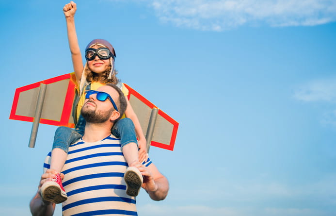 A young father with his pre-teen daughter on his shoulders; she is wearing wings and goggles